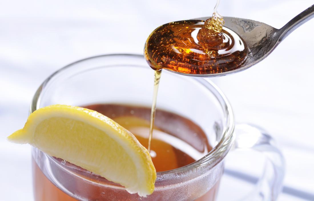 Honey being drizzled onto spoon and into mug of tea with lemon slice.